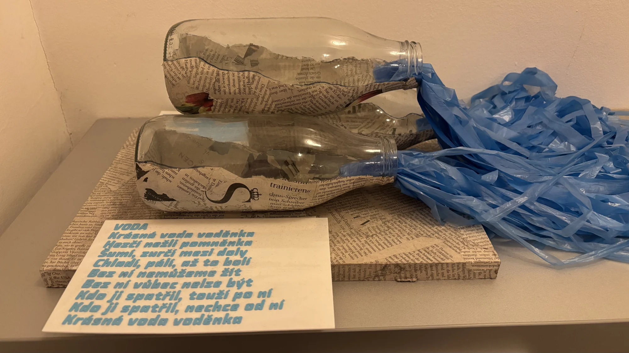 The Water artwork displayed with glass bottles, newspaper collage, blue ribbons and Magdaléna’s poem